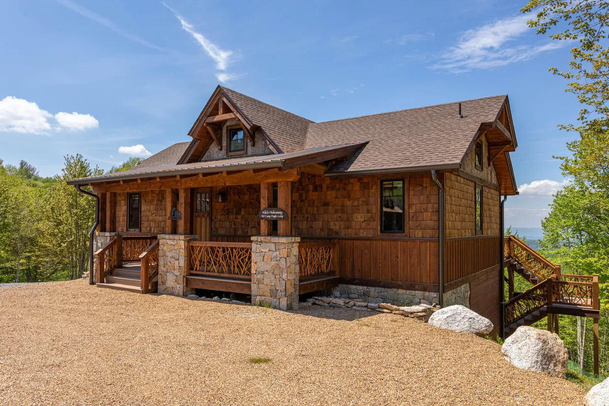 This image shows the home's wooden facade with a welcoming front porch, landscaped path, and stone accents. The scene is enveloped by natural greenery, suggesting tranquility and opportunities for outdoor relaxation or gathering.