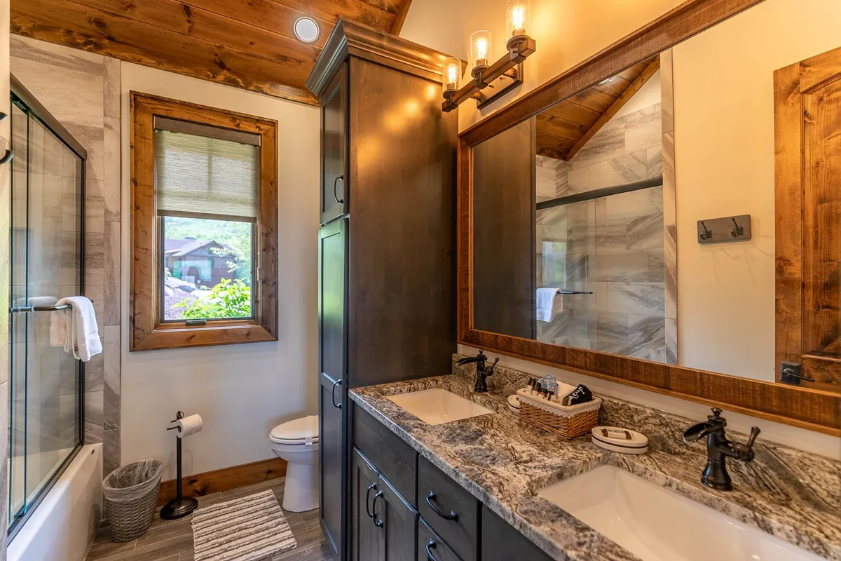 A spacious en-suite bathroom with a double sink vanity, granite countertops, a large framed mirror, a bathtub, and a window overlooking greenery. The space is bright, calm, and perfect for relaxing routines.