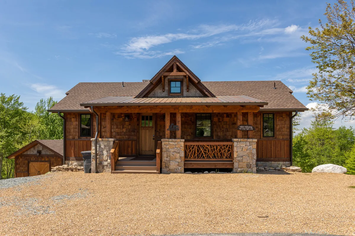 The front porch of the home features wooden rocking chairs and stone accents, inviting guests to relax and enjoy the views. The exterior architecture is warm and welcoming, with natural materials blending into the surrounding landscape. The porch overlooks greenery, hinting at the connection between the home and the outdoors.
