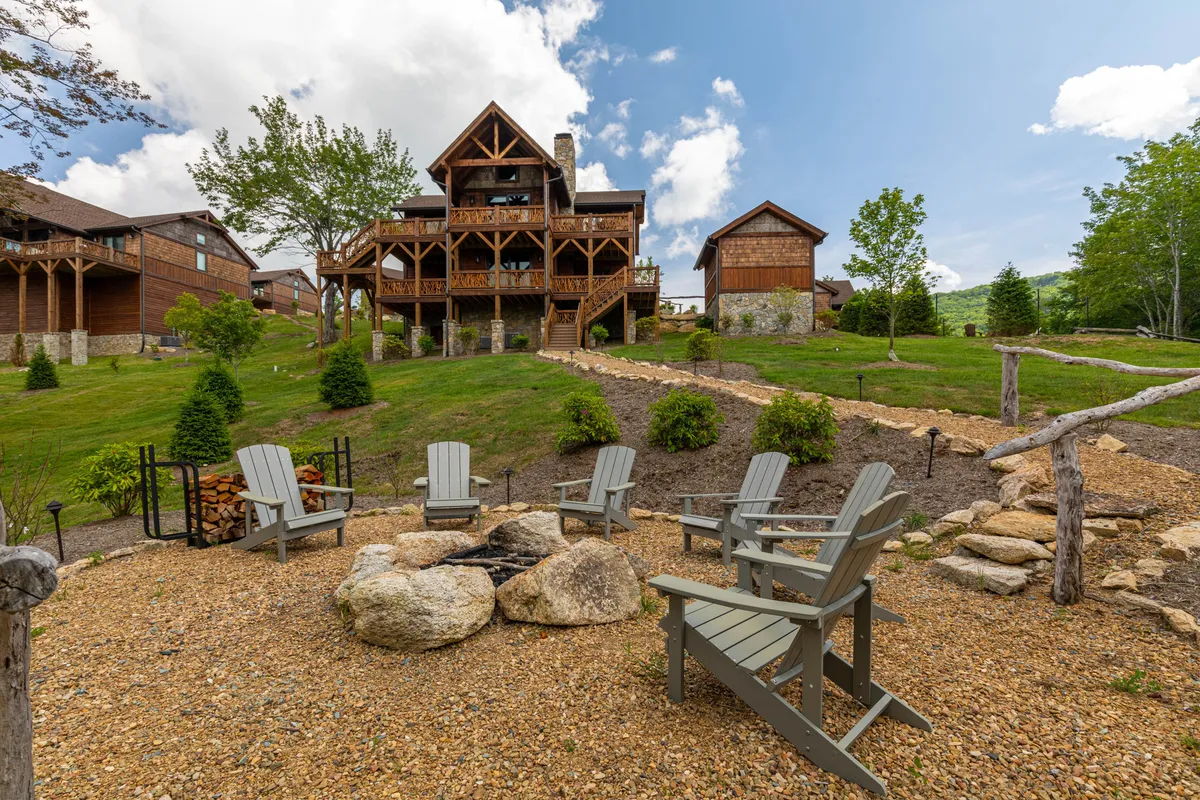 A wide exterior view featuring a large wooden house, a lawn seating area, and a prominent fire pit surrounded by chairs. The scene radiates a sense of gathering and connection with nature, set against a tranquil mountain backdrop.