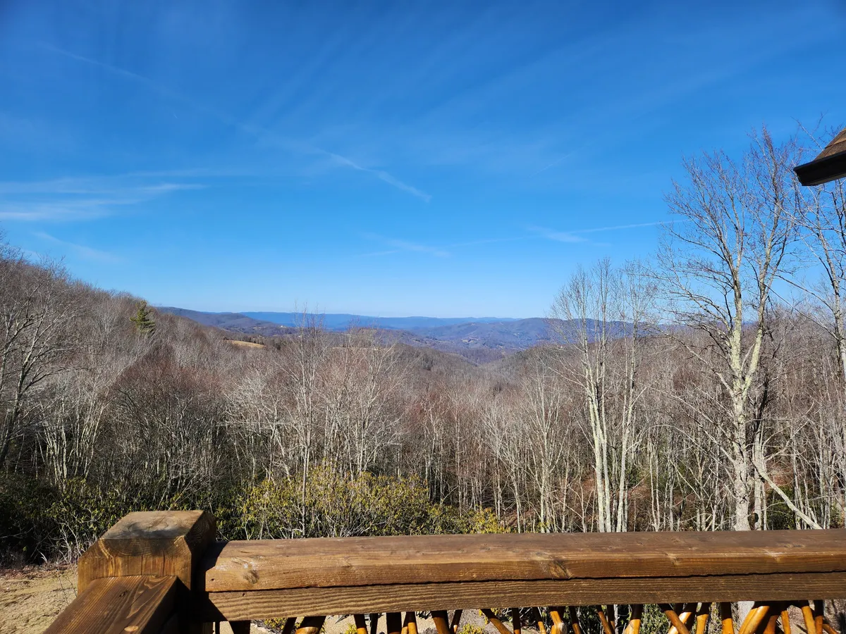 A tranquil mountain landscape with bare trees, rolling hills, and a brilliant blue sky. The scene evokes the peace and beauty of North Carolina's mountain setting, with space for activities like hiking or stargazing. The deck setting alludes to morning coffee or relaxing evenings outdoors.