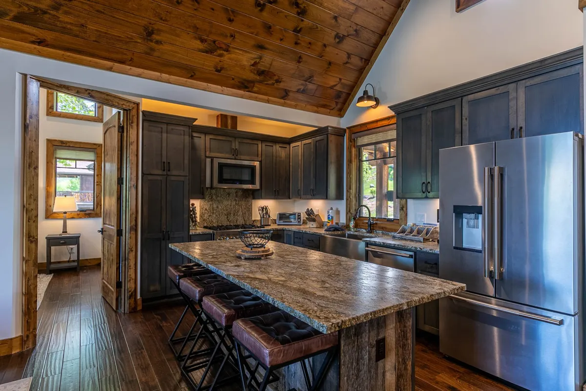 A warm kitchen scene with a granite countertop island, wood cabinetry, stainless steel appliances, and high ceilings with exposed beams. The layout is ideal for group meal prep and casual conversations around the island.
