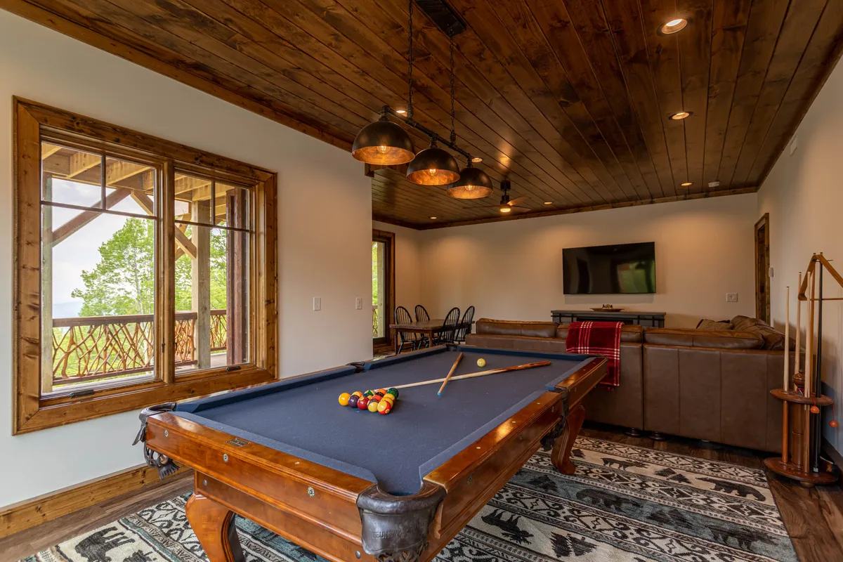 This image captures a vibrant game room featuring a pool table as the centerpiece, large windows letting in ample natural light, a comfortable sofa, and a visible dining area to the side. The room is inviting and set up for both recreation and relaxation, perfectly echoing the lower-level retreat's social atmosphere.
