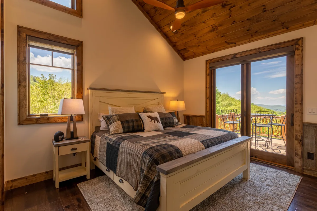 A cozy king bedroom featuring a bed with plaid bedding, large windows that let in natural light, and wooden furniture. The windows offer a glimpse of the outdoors, reinforcing a sense of tranquility and connection to the scenic setting.