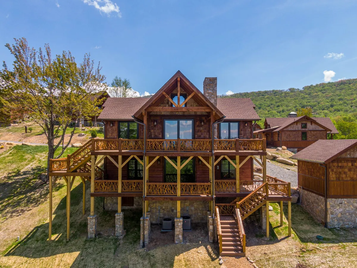 The image showcases the inviting exterior of a single-family mountain home nestled among lush greenery. Prominent features include expansive windows that flood the interior with light, a spacious wooden deck with an ornate railing perfect for gatherings, a sturdy stone foundation, and panoramic views of the rolling hills—ideal for soaking in sunsets or morning coffee in nature's embrace.