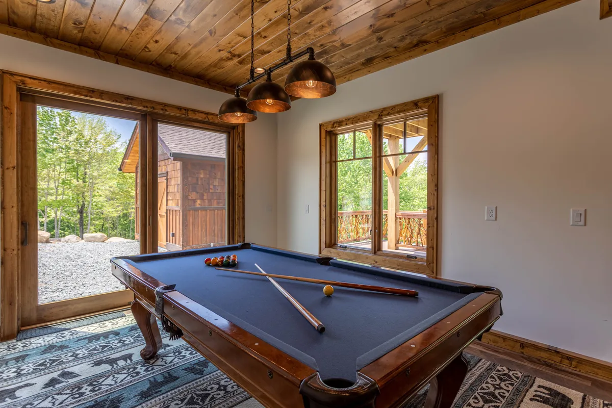 An inviting interior game room featuring a pool table, large windows with scenic views, a warm wooden ceiling, and stylish pendant lights. The space captures a playful, social vibe perfect for family gatherings and game nights.