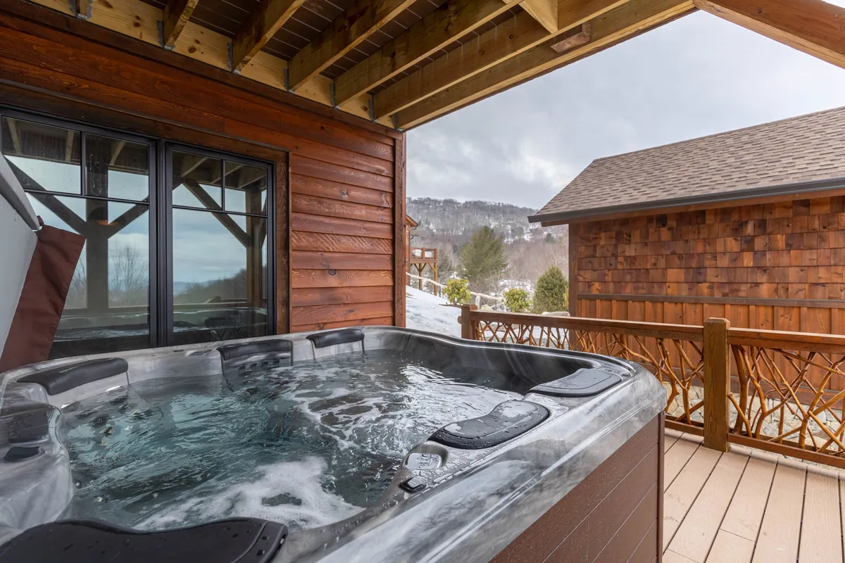 A tranquil exterior shot showing a wooden deck with a bubbling hot tub overlooking a sweeping mountain view. The space is open to the sky, inviting relaxation and socializing amidst scenic natural surroundings.